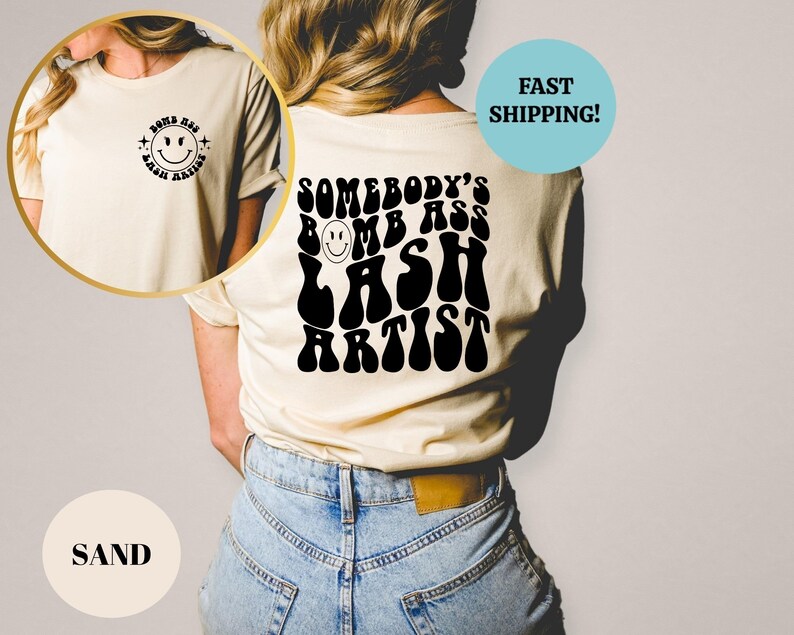 May include: A white t-shirt with a black graphic that says "Somebody's Bomb Ass Lash Artist" in a retro font. The graphic includes a smiley face.