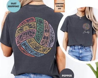 May include: A charcoal gray short-sleeve t-shirt featuring a large, circular volleyball graphic on the back filled with colorful floral and leaf patterns. The front left chest displays a smaller version of the same volleyball design with the name Emily written in white cursive script below it.