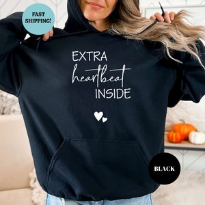 Pode incluir: Camisola de moletom com capuz preta com o texto "EXTRA heartbeat INSIDE" e dois pequenos cora&ccedil;&otilde;es brancos impressos na frente.