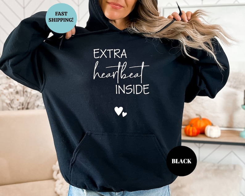 Pode incluir: Camisola de moletom com capuz preta com o texto "EXTRA heartbeat INSIDE" e dois pequenos cora&ccedil;&otilde;es brancos impressos na frente.