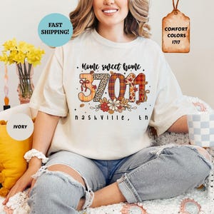 May include: An ivory t-shirt featuring a graphic print with the text Home sweet home and Nashville, TN. The numbers 37011 are displayed in a large font with a mix of leopard print, plaid, and autumn leaf patterns, accented by small floral illustrations.