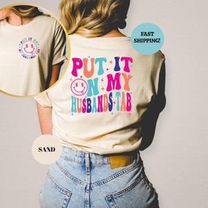 May include: A light beige t-shirt with a colorful graphic that says "Put it on my husband's tab" with a smiley face and dollar signs. The shirt is being modeled by a person wearing blue jeans.
