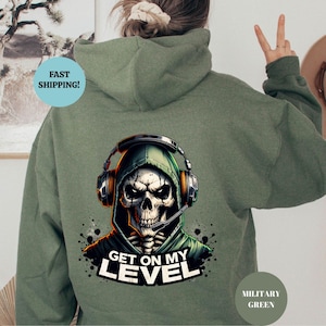 May include: Military green hoodie featuring a skull wearing a headset and a green hood. The graphic includes the text "GET ON MY LEVEL" in white. The hoodie is a solid color with a drawstring hood.