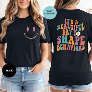 以下が含まれることがあります： 黒いTシャツ。前面にはスマイリーフェイスと虹色のフォントで「BEHAVIOR SQUAD」の文字。背面には「IT'S A BEAUTIFUL DAY TO SHAPE BEHAVIORS」の文字とデイジーのグラフィック。
