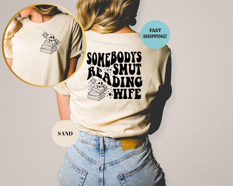 May include: A beige t-shirt with the text "Somebody's Smut Reading Wife" in black, with a skull and stack of books graphic. The shirt is being worn by a person with blue jeans.