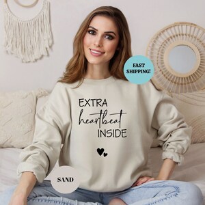 Pode incluir: Uma camisa de moletom bege claro com o texto "EXTRA heartbeat INSIDE" e dois pequenos cora&ccedil;&otilde;es pretos impressos na frente. A modelo est&aacute; vestindo a camisa de moletom e sentada em uma cama.