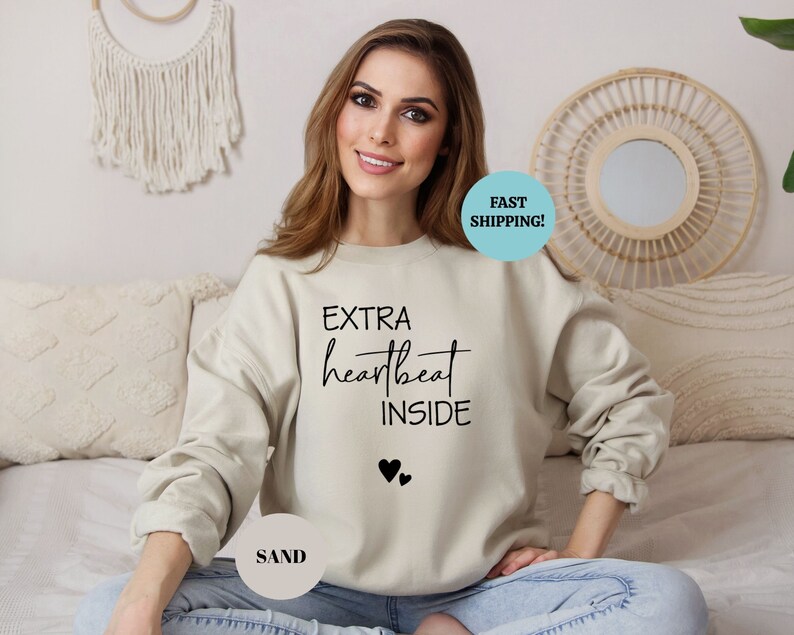 Pode incluir: Uma camisa de moletom bege claro com o texto "EXTRA heartbeat INSIDE" e dois pequenos cora&ccedil;&otilde;es pretos impressos na frente. A modelo est&aacute; vestindo a camisa de moletom e sentada em uma cama.