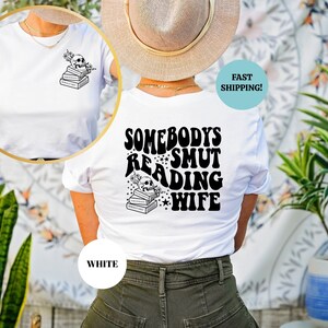 May include: White t-shirt with a black graphic on the back that says "Somebody's Smut Reading Wife" with a skull and stack of books. The front of the shirt has a smaller graphic of a skull and stack of books.