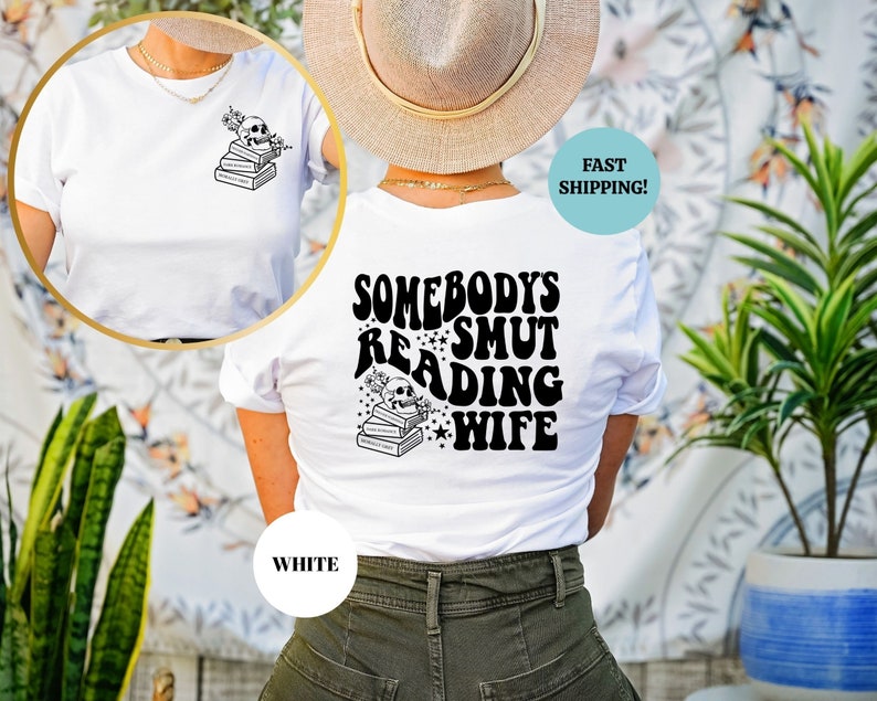 May include: White t-shirt with a black graphic on the back that says "Somebody's Smut Reading Wife" with a skull and stack of books. The front of the shirt has a smaller graphic of a skull and stack of books.