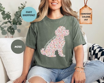 May include: A moss green short-sleeve t-shirt featuring a graphic of a dog silhouette filled with a colourful floral pattern. The text "GOLDENDOODLE MOM" is written in a script font across the dog's body. The shirt has a crew neckline.