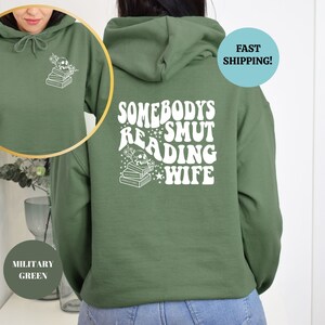 May include: A green hooded sweatshirt with the text "Somebody's Smut Reading Wife" printed on the back in white. The text is in a retro font style with stars and a stack of books.