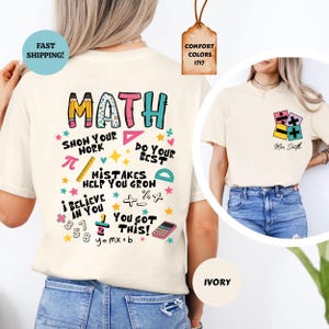 May include: Ivory-colored t-shirt with a colorful math-themed design. The back features the word "MATH" and encouraging phrases like "Show Your Work" and "You Got This!" A smaller design with math symbols is on the front.