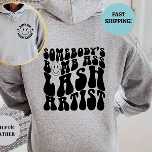 May include: Gray hoodie with a black graphic that says "Somebody's Bomb Ass Lash Artist" in a retro font. The graphic includes a smiley face.