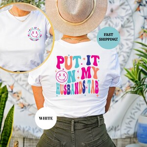 May include: White t-shirt with a colorful graphic that says "Put it on my husband's tab" with a smiley face and dollar signs. The shirt is shown on a model wearing it.