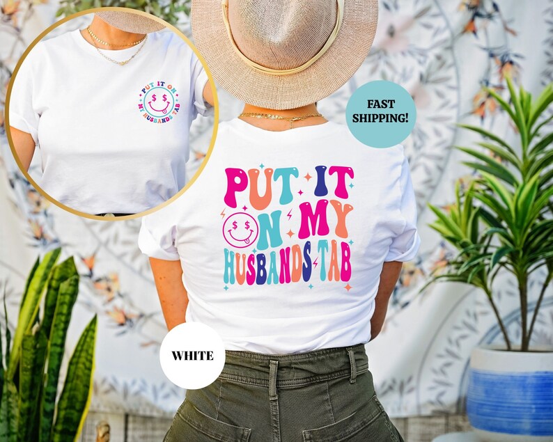 May include: White t-shirt with a colorful graphic that says "Put it on my husband's tab" with a smiley face and dollar signs. The shirt is shown on a model wearing it.
