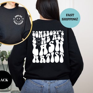 May include: Black sweatshirt with white text that reads "Somebody's Bomb Ass Lash Artist" in a retro font. The text is centered on the back of the sweatshirt. A smiley face is included in the text.