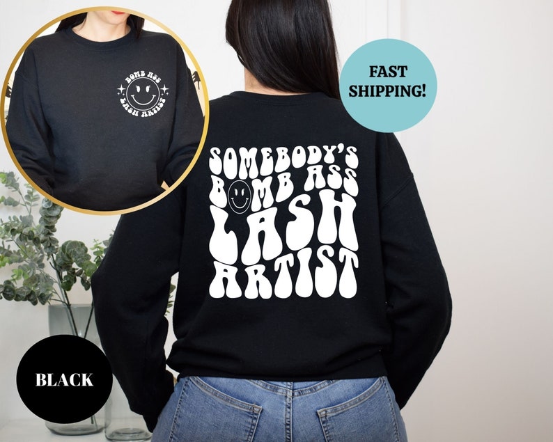 May include: Black sweatshirt with white text that reads "Somebody's Bomb Ass Lash Artist" in a retro font. The text is centered on the back of the sweatshirt. A smiley face is included in the text.