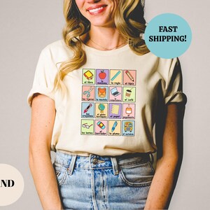 May include: A beige t-shirt with a graphic of a grid of colorful squares featuring Spanish words and illustrations of school supplies. The words include "el libro", "la regla", "el l&aacute;piz", "las tijeras", "el machie", "la mochila", "los crayones", "el globo terrestre", "el papel", "los lentes", "el borrador", "la pluma", "el autob&uacute;s", and "el caf&eacute;".