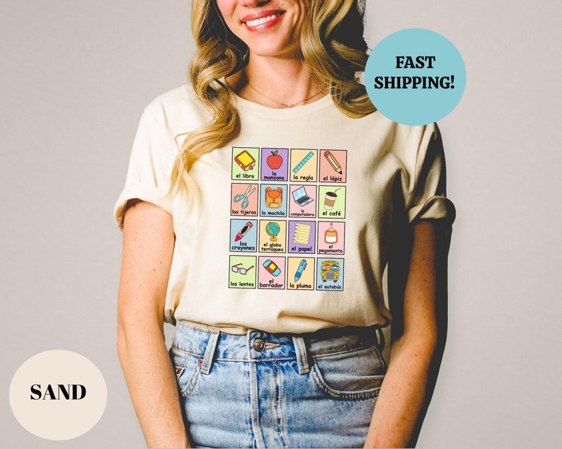 May include: A beige t-shirt with a graphic of a grid of colorful squares featuring Spanish words and illustrations of school supplies. The words include "el libro", "la regla", "el l&aacute;piz", "las tijeras", "el machie", "la mochila", "los crayones", "el globo terrestre", "el papel", "los lentes", "el borrador", "la pluma", "el autob&uacute;s", and "el caf&eacute;".
