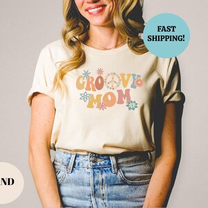 May include: A woman wearing a sand colored t-shirt with the text "Groovy Mom" in retro font and colorful flowers.