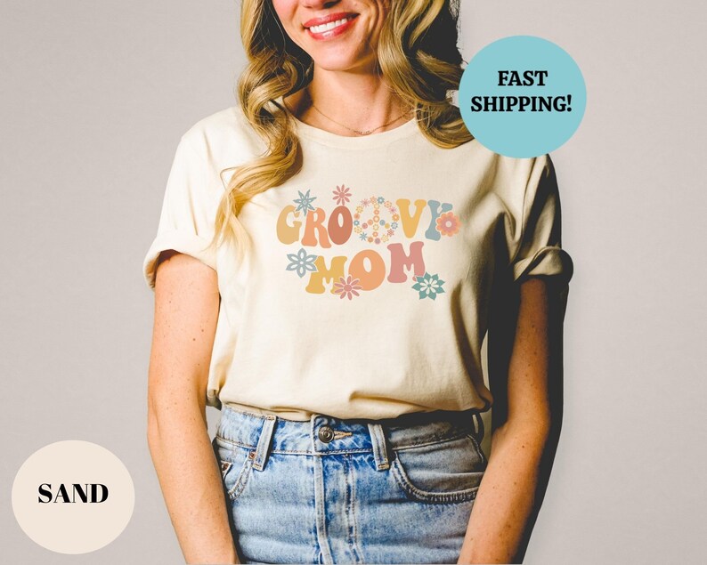May include: A woman wearing a sand colored t-shirt with the text "Groovy Mom" in retro font and colorful flowers.