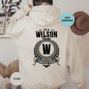 May include: A cream-colored hooded sweatshirt with black text that reads "It's a Wilson thing. You wouldn't understand." The text is surrounded by a black wreath with a black "W" in the center.