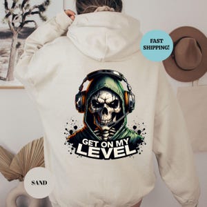 May include: Sand-colored hoodie featuring a graphic of a skull wearing a headset and a green hooded jacket. The text "GET ON MY LEVEL" is below the skull. The hoodie is a casual garment.
