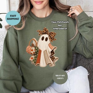 Puede incluir: Una sudadera de cuello redondo color verde militar con un gráfico central de un fantasma que lleva un lazo con estampado de leopardo y sostiene una cesta de calabazas. El diseño muestra un patrón de patchwork con texturas otoñales.