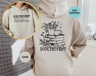 May include: A beige hoodie with a black graphic design on the back. The design features a stack of books, a cup of coffee, flowers, and the text "BOOKTROVERT". The front of the hoodie has the text "BOOKTROVERT" with a definition.