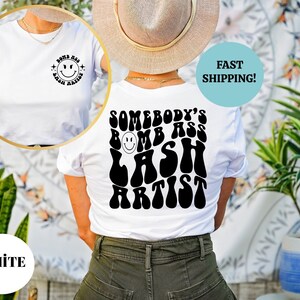 May include: White t-shirt with a black graphic on the back that says "Somebody's Bomb Ass Lash Artist" in a retro font. The graphic features a smiley face.