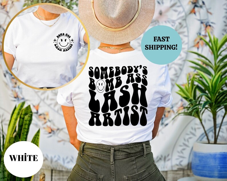 May include: White t-shirt with a black graphic on the back that says "Somebody's Bomb Ass Lash Artist" in a retro font. The graphic features a smiley face.