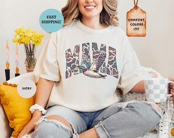May include: An ivory short-sleeve t-shirt featuring the word MAMA in a camouflage pattern with a mallard duck graphic flying across the letters. The shirt is labeled as Comfort Colors 1717 and is shown in the color ivory.
