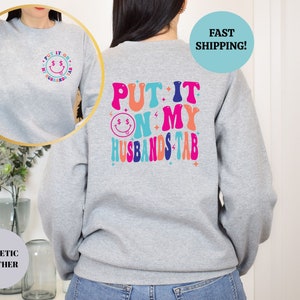 May include: Gray sweatshirt with the text "PUT IT ON MY HUSBANDS TAB" in colorful retro letters on the back. The front of the sweatshirt has a small smiley face with dollar signs for eyes.