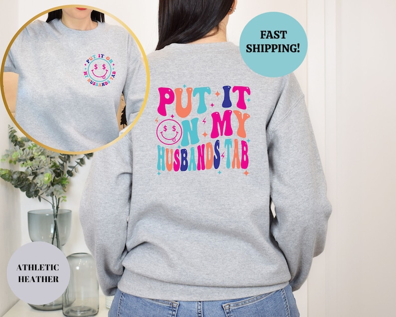 May include: Gray sweatshirt with the text "PUT IT ON MY HUSBANDS TAB" in colorful retro letters on the back. The front of the sweatshirt has a small smiley face with dollar signs for eyes.