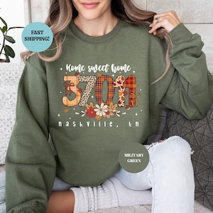 May include: Military green sweatshirt with the text "Home sweet home" and "37011 Nashville, TN" in a decorative font. The numbers are patterned with animal print and plaid. The sweatshirt is a solid color.