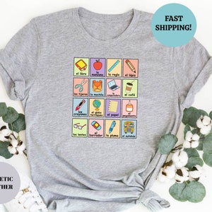 May include: A heather grey t-shirt with a colorful graphic of Spanish school supplies and the words "el libro", "la manzana", "la regla", "el l&aacute;piz", "las tijeras", "la mochila", "la computadora", "el caf&eacute;", "los crayones", "el globo terraqueo", "el papel", "el pegamento", "los lentes", "el borrador", "la pluma", "el autob&uacute;s".