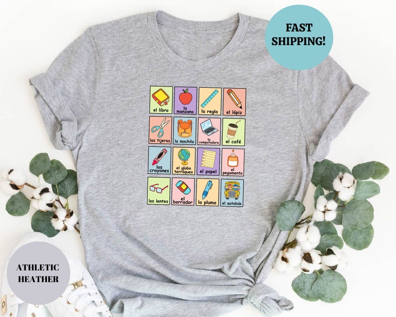 May include: A heather grey t-shirt with a colorful graphic of Spanish school supplies and the words "el libro", "la manzana", "la regla", "el l&aacute;piz", "las tijeras", "la mochila", "la computadora", "el caf&eacute;", "los crayones", "el globo terraqueo", "el papel", "el pegamento", "los lentes", "el borrador", "la pluma", "el autob&uacute;s".