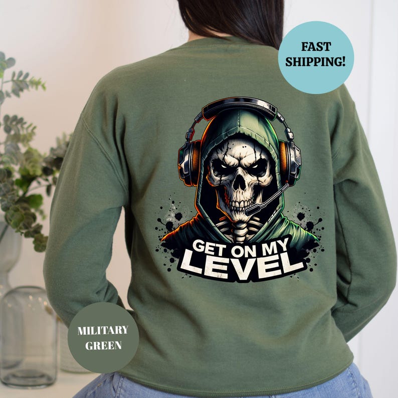 May include: Military green sweatshirt featuring a graphic of a skull wearing a headset and hooded sweatshirt. The text "GET ON MY LEVEL" is below the skull. The sweatshirt is a solid color.