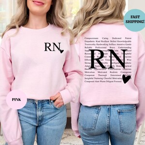 May include: Pink sweatshirt with the letters "RN" and a heart on the front. The back of the sweatshirt has the letters "RN" in a large font surrounded by a word cloud of positive adjectives describing a nurse.