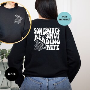 May include: Black sweatshirt with a white graphic on the back that says "Somebody's Smut Reading Wife" with a skull and books. The front of the sweatshirt has a small graphic of a skull and books.