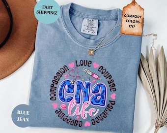 May include: A blue jean colored t-shirt featuring a graphic design with the text CNA life in blue leopard print and pink script. The design includes a stethoscope, a syringe, pink hearts, and the words compassion, love, courage, and patience.