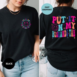 May include: Black t-shirt with a colorful graphic that says "Put it on my husband's tab" with a smiley face and dollar signs.