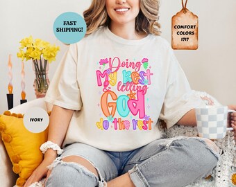May include: An ivory short-sleeve t-shirt featuring a colorful graphic print with the text "Doing My Best & letting God do the rest" in pink, orange, and blue bubble letters, accented with small flower and star illustrations.