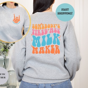 Pu&ograve; includere: Felpa grigia con una grafica in stile retr&ograve; sul retro che dice "Somebody's Tired Ass Milk Maker" in rosa, blu e arancione. La parte anteriore della felpa ha una piccola grafica di una mano scheletrica che fa il gesto rock and roll.