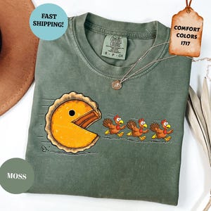May include: Moss green Comfort Colors t-shirt featuring a pie-shaped character chasing three cartoon turkeys. The shirt has a crew neck and short sleeves. The tag reads "Comfort Colors 1717".