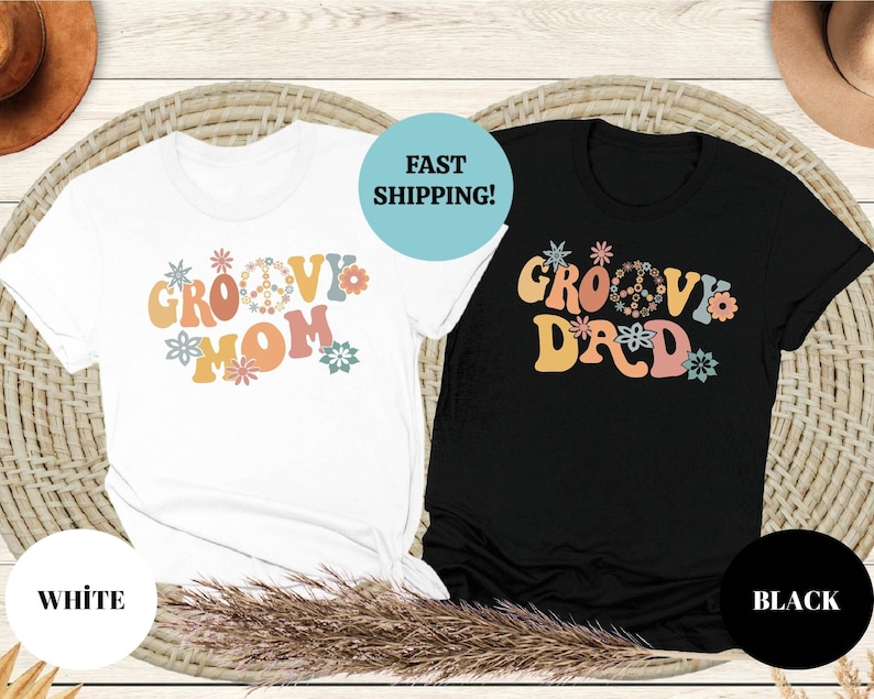 May include: Two t-shirts with a groovy font design. One shirt is white with the text "Groovy Mom" and the other shirt is black with the text "Groovy Dad". Both shirts feature a peace sign and flowers.