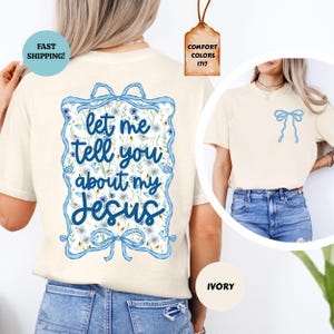May include: Ivory-colored t-shirt with a blue bow graphic on the front and a larger design on the back. The back design features the text "let me tell you about my Jesus" in a blue frame with floral accents. The shirt has short sleeves.