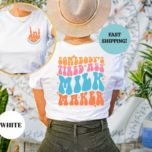 Pu&ograve; includere: Maglietta bianca con una grafica sul retro che dice "Somebody's Tired Ass Milk Maker" in un font retr&ograve; con colori rosa, blu e arancio. La parte anteriore della maglietta ha un piccolo grafico di una mano con il dito medio esteso.