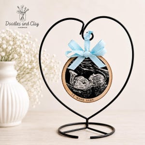 May include: A wooden disc ornament featuring a baby sonogram image, adorned with a light blue ribbon. It's presented in a black metal heart-shaped stand. The text "12 week scan" is inscribed on the ornament.