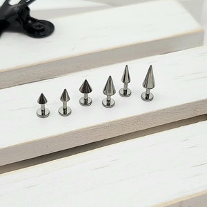 May include: A set of six silver-toned surgical steel spike earrings in various sizes. The earrings are arranged in order of size, with the smallest on the left and the largest on the right.